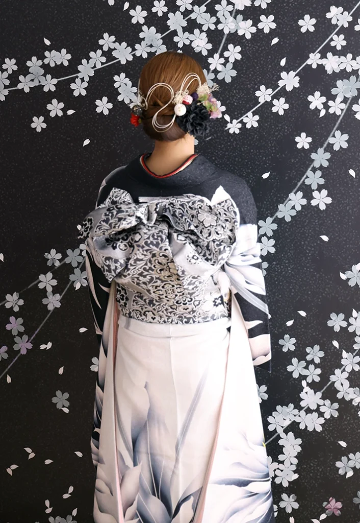 A-24 白銀に大花の振袖後ろ姿｜黒い桜模様の背景にレース柄の帯結びが映えるスタジオ撮影カット